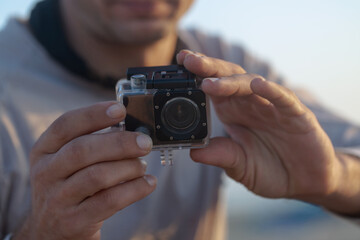 Small modern action camera in waterproof housing filming in shallow water on beach, man holding recording equipment sitting outdoors. Selective focus