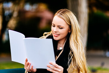 Young woman sitting on a park bench reading a magazine with a blank cover page. Magazine as mock-up
