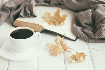 Naklejka na ściany i meble Autumn leaves, cup of coffee, warm wool sweater with open old book on white rustic wooden boards. Cozy home reading, hygge concept. Shallow depth of field