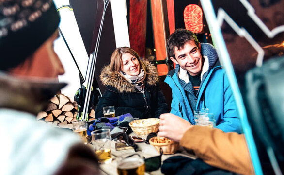 Happy friends drinking beer and eating chips at apreski outdoors - Young people having fun together at night bar restaurant after skiing with snow equipment - Friendship concept with focus on woman