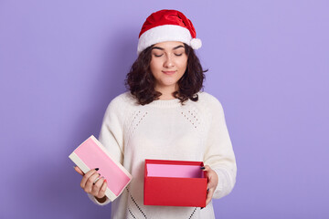 Brunette woman in santa claus hat with opened gift box in hands, looking directly at camera, wearing white warm sweater, standing isolated over lilac background.