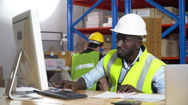 african warehouse manager sitting at desk using computer check inventory items online at distribution warehouse. black man worker  in Hard Hat typing keyboard entering data document  in workplace