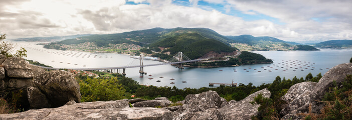 Panorama view of the Ria de Vigo estuary from Redondela on a cloudy Summer afternoon, with the recently extended Rande bridge on the center and the Atlantic Ocean on the left. Long exposure. - obrazy, fototapety, plakaty