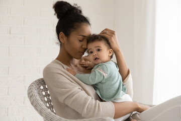 Happy African American mother hold hug newborn small biracial daughter shown love and care. Smiling ethnic mom embrace cuddle toddler baby child relax in chair at home together. Childcare concept.