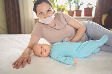 Obraz na płótnie Canvas Young mother in protective face mask with newborn cute infant baby in blue jumpsuit lying on bed