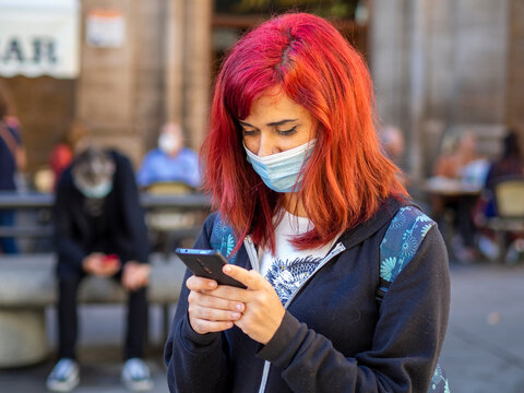Mujer joven con pelo rojo usando su teléfono móvil vistiendo mascarilla para respetar las medidas de seguridad y distanciamiento social de la pandemia del coronavirus