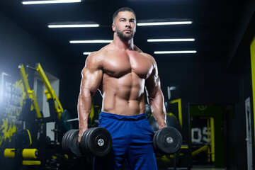 Plakat Front view of a muscular bodybuilder posing with dumbbells in blue shorts in a gym
