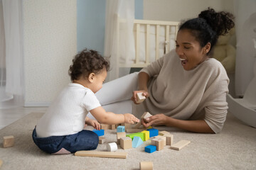 Smiling african American young mom lying on floor at home kids room play with building blocks with little baby toddler. Happy loving caring biracial mother engaged in funny activity with infant child.