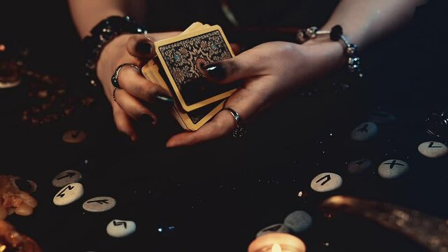 ivination and magic. A fortune teller shuffles Tarot cards. Close-up of hands with dark manicure and rings. A black table with scattered runes