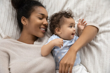 Close up top view of loving young African American mom lying in comfortable bed hug cuddle small cute toddler baby. Smiling caring biracial mother awaken in home bedroom with little newborn child.