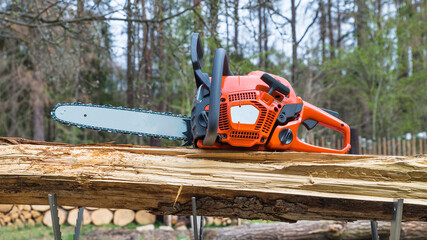 Orange hand gasoline chainsaw on wooden log in metal sawhorse. Professional power saw with black...