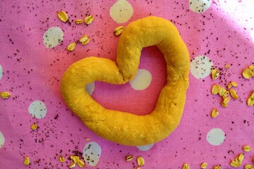 Heart shaped bread on a pink tablecloth with white polka dots surrounded by rolled oats and poppy seeds