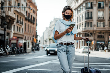 Young traveling woman with suitcase on a sunny city street. Traveler wearing mask on vacation.