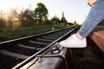 The old suitcase stands right next to the railway tracks on the platform and on it are the feet in the sports shoes of the young traveler.