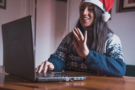 Una chica joven haciendo una video llamada saluda a su ordenador portátil mientras lleva puesto un gorro de Santa Claus - Powered by Adobe