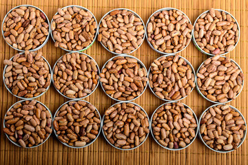 Top view of fifteen round bowls with many fresh organic red beans on a wooden table in warm light, brown monochrome indoor background photographed with selective focus.