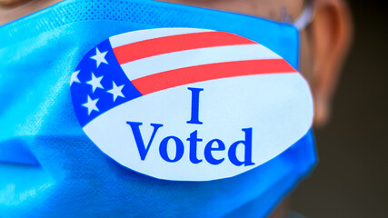 Voting during Coronavirus Pandemic concept. Man puts I Voted sticker with American Flag on the face mask.