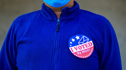 Man in mask from minority group produly holds Voted By Mail sticker during US Election closeup.