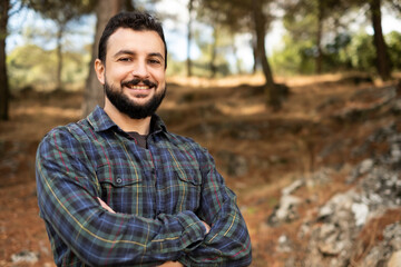 Man with beard and plaid shirt of aesthetic lumberjack crosses his arms in the middle of nature in portrait looking at camera