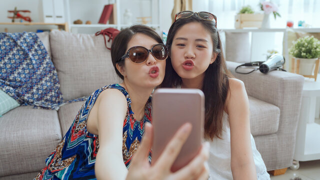 Female best friends sitting on floor in living room apartment and taking selfies by cellphone in summer break