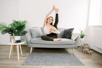 Pretty young Caucasian yoga fit woman sitting on gray sofa and posing to camera, stretching leg up, smiling and holding her smartphone. Woman watching online video yoga lessons