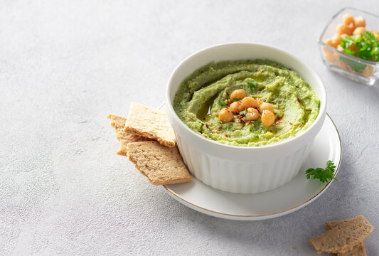 white bowl with avocado hummus with chickpeas, parsley, spices and crackers on gray background. Healthy vegan snack. side view. place for text.