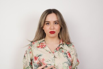 Waist up shot of joyful young woman looking to the camera, thinking or wondering about something. Both arms down, neutral facial expression.