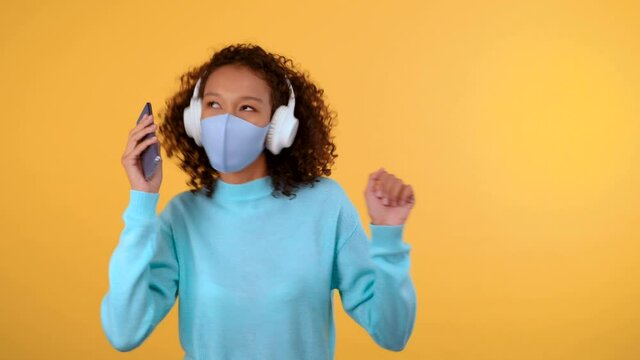 cute funny young woman with afro hair dancing on an isolated yellow background she is in her protective medical mask. A woman holds her smartphone in her hand. 4K