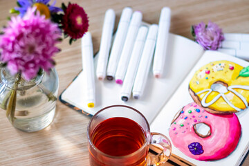 Transparent cup of tea on sketching donuts background. All placed on wooden table with transparent cup of tea, notebook, markers, and flowers. Hobby. Drawing donuts.