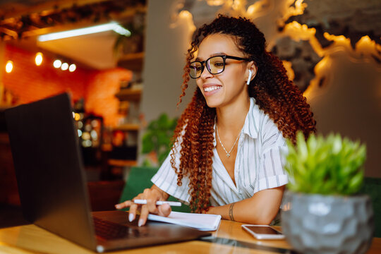 African American woman wearing headphones calling on laptop, talk by webcam, video conference sitting at cafe. Online meeting in video call with friends, relative or business partners.