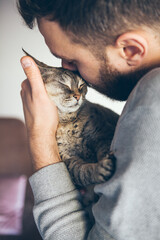 Portrait of happy cat with closed eyes and beard man. Handsome young animal-lover man hugs and cuddles his tabby Devon Rex cat. Sweet and mellow kitty. Breed with hypoallergenic fur, no shedding.