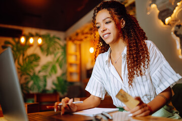 African American woman with credit card and laptop computer doing online shopping sitting at cafe or restaurant. Online shopping, e-commerce, internet banking, spending money.