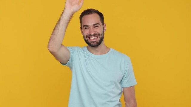 Handsome happy young man guy 20s years old in basic casual blue t-shirt isolated on yellow background studio. People lifestyle concept. Looking camera waving and greeting with hand as notices someone