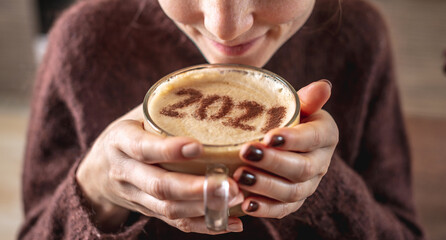 A young woman weared in a cozy warm sweater is enjoying a hot coffee. There are numbers 2021 on the milk foam. The concept of New year