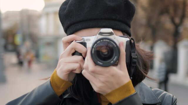 Young woman photograph with photo camera looking straight, doing pictures close up. Beautiful asian woman in black beret holding a camera shooting in the street