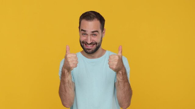 Bearded young man 20s in basic casual blue t-shirt isolated on yellow background studio. People lifestyle concept. Looking camera charming smile pointing finger on you say cool approval thumbs up like