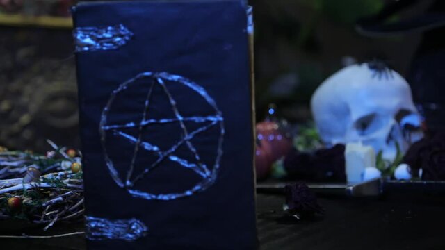Close-up, change of focus from human skull to black book with pentagram, camera smooth movement. The concept of a witch's table for rituals on the eve of halloween.