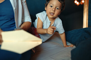 Horizontal image of adorable active little boy playing in bedroom, pointing finger at camera, his unrecognizable mother in foreground folding paper, making origami toy for him. Selective focus