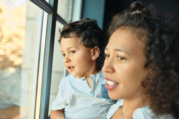 Family, togetherness, leisure and relaxation. Young mixed race female with little son in her arms having curious facial expression looking outside through window. Selective focus on boy face