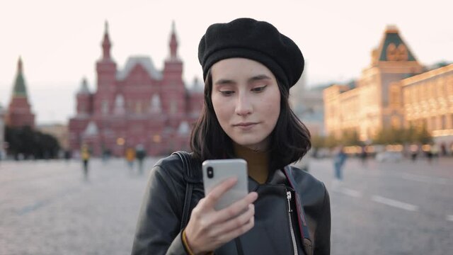 Young smiling girl on Red Square using phone, looking at the screen. Young asian woman wearing beret and leather jacket standing on background of museum and GUM