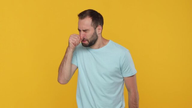 Bearded young guy 20s in blue t-shirt isolated on yellow background studio. Healthy lifestyle ill sick disease treatment cold season concept. Looking at camera feeling bad sneezing in hands allergy