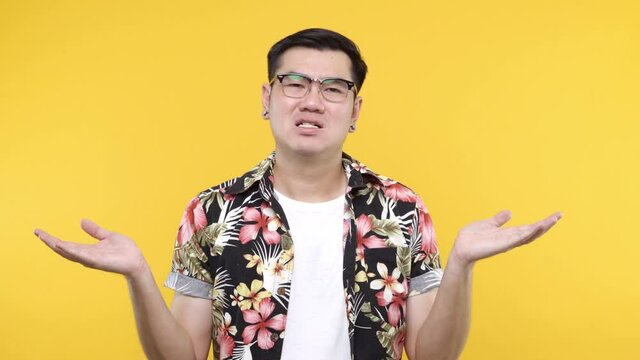 A serious young asian man wearing aloha shirt saying NO while crossing his hands standing isolated on yellow background. 4k resolution.