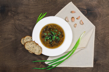 Vegetarian soup with wild white mushrooms, vegetables, green onions and healthy cereal bread. Traditional dish in a white plate on a dark wooden table background with a textile napkin, top view