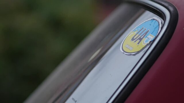 UA sticker in the blue and yellow colors of the national Ukrainian flag on the rear window of a car. A vintage car stands on the road. Transport passes by. Green foliage in the background
