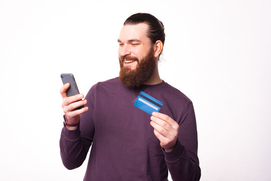 Photo of a bearded man looking in his phone and is holding a credit card .