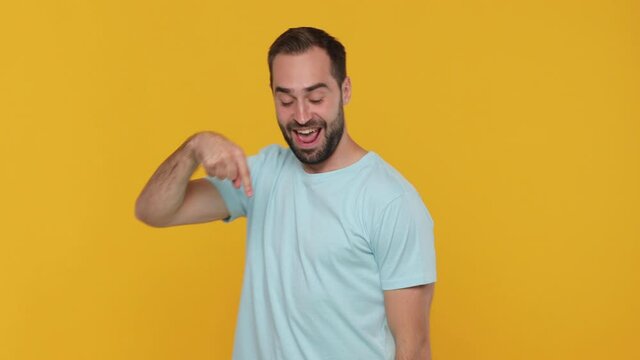Fun expressive young man 20s years old in basic casual blue t-shirt look camera pointing fingers hands down on copy space workspace area isolated on yellow background studio. People lifestyle concept