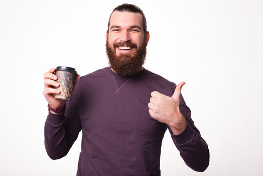 A picture of a young bearded man is smiling at the camera and holding a cup of hot drink is showing a thumb up .