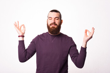 A young bearded man is sitting in relaxing pose near a white wall .