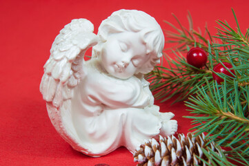 figurine of a Christmas angel sitting near the branches of a Christmas tree with cones on a red background