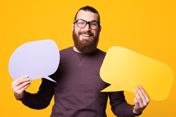 Picture of a man with beard wearing glasses is holding two speech bubbles smiling at the camera .